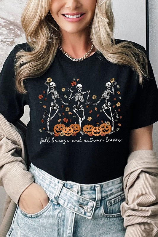 A person wearing a high-quality, unisex dark grey "Fall Breeze and Autumn Leaves Skeleton Graphic Tee" featuring dancing skeletons, pumpkins, and autumn leaves, holding an orange mug. They are dressed in a knitted cardigan and ripped jeans. The text on the shirt reads "Fall breeze and autumn leaves.