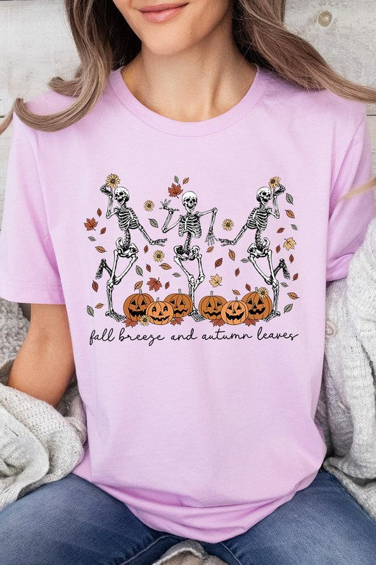 A person wearing a high-quality, unisex dark grey "Fall Breeze and Autumn Leaves Skeleton Graphic Tee" featuring dancing skeletons, pumpkins, and autumn leaves, holding an orange mug. They are dressed in a knitted cardigan and ripped jeans. The text on the shirt reads "Fall breeze and autumn leaves.