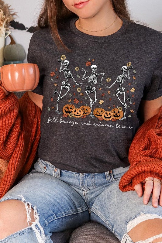 A person wearing a high-quality, unisex dark grey "Fall Breeze and Autumn Leaves Skeleton Graphic Tee" featuring dancing skeletons, pumpkins, and autumn leaves, holding an orange mug. They are dressed in a knitted cardigan and ripped jeans. The text on the shirt reads "Fall breeze and autumn leaves.