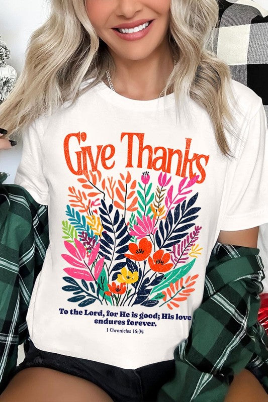 A person wears the Give Thanks Christian Graphic Tee, a high-quality printed design white t-shirt featuring colorful floral graphics and the text "Give Thanks. To the Lord, for He is good; His love endures forever. 1 Chronicles 16:34." Perfect as a family group uniform or a Christian graphic tee for gatherings.