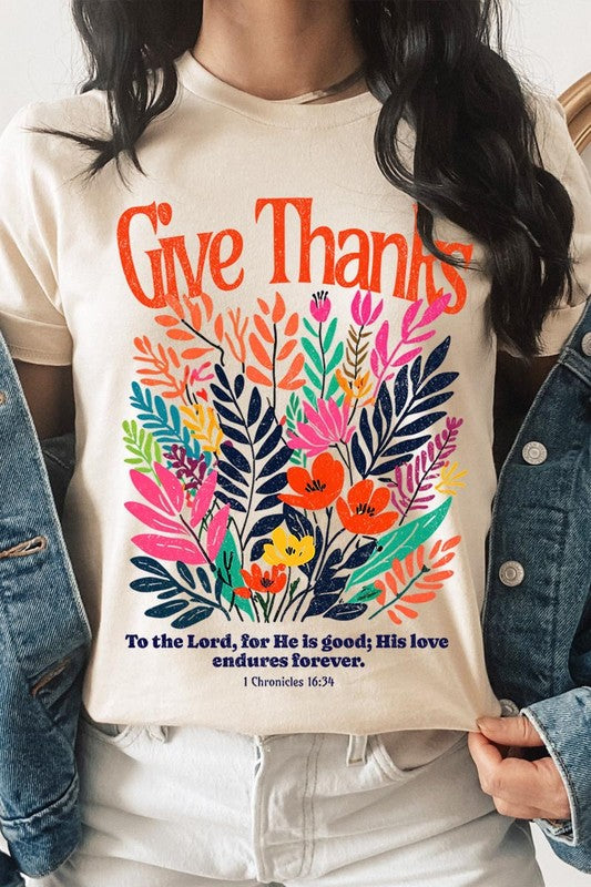 A person wears the Give Thanks Christian Graphic Tee, a high-quality printed design white t-shirt featuring colorful floral graphics and the text "Give Thanks. To the Lord, for He is good; His love endures forever. 1 Chronicles 16:34." Perfect as a family group uniform or a Christian graphic tee for gatherings.