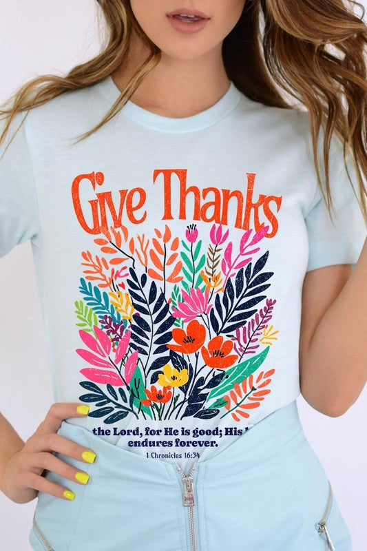 A person wears the Give Thanks Christian Graphic Tee, a high-quality printed design white t-shirt featuring colorful floral graphics and the text "Give Thanks. To the Lord, for He is good; His love endures forever. 1 Chronicles 16:34." Perfect as a family group uniform or a Christian graphic tee for gatherings.