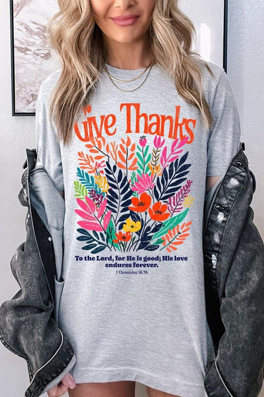 A person wears the Give Thanks Christian Graphic Tee, a high-quality printed design white t-shirt featuring colorful floral graphics and the text "Give Thanks. To the Lord, for He is good; His love endures forever. 1 Chronicles 16:34." Perfect as a family group uniform or a Christian graphic tee for gatherings.