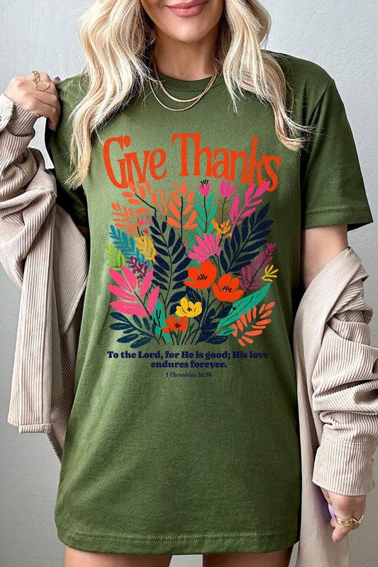 A person wears the Give Thanks Christian Graphic Tee, a high-quality printed design white t-shirt featuring colorful floral graphics and the text "Give Thanks. To the Lord, for He is good; His love endures forever. 1 Chronicles 16:34." Perfect as a family group uniform or a Christian graphic tee for gatherings.