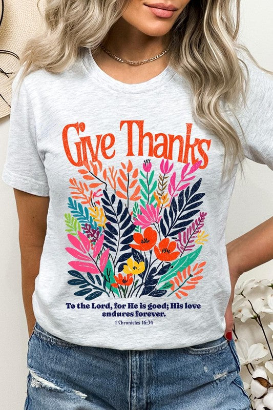 A person wears the Give Thanks Christian Graphic Tee, a high-quality printed design white t-shirt featuring colorful floral graphics and the text "Give Thanks. To the Lord, for He is good; His love endures forever. 1 Chronicles 16:34." Perfect as a family group uniform or a Christian graphic tee for gatherings.