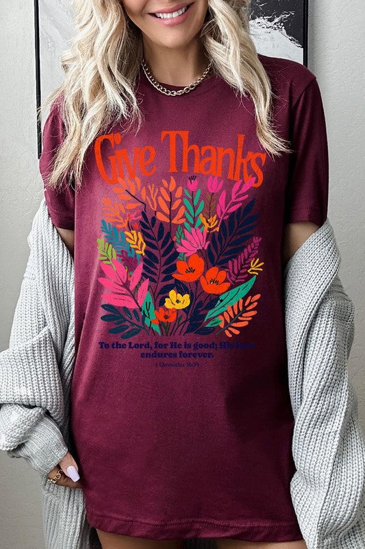 A person wears the Give Thanks Christian Graphic Tee, a high-quality printed design white t-shirt featuring colorful floral graphics and the text "Give Thanks. To the Lord, for He is good; His love endures forever. 1 Chronicles 16:34." Perfect as a family group uniform or a Christian graphic tee for gatherings.