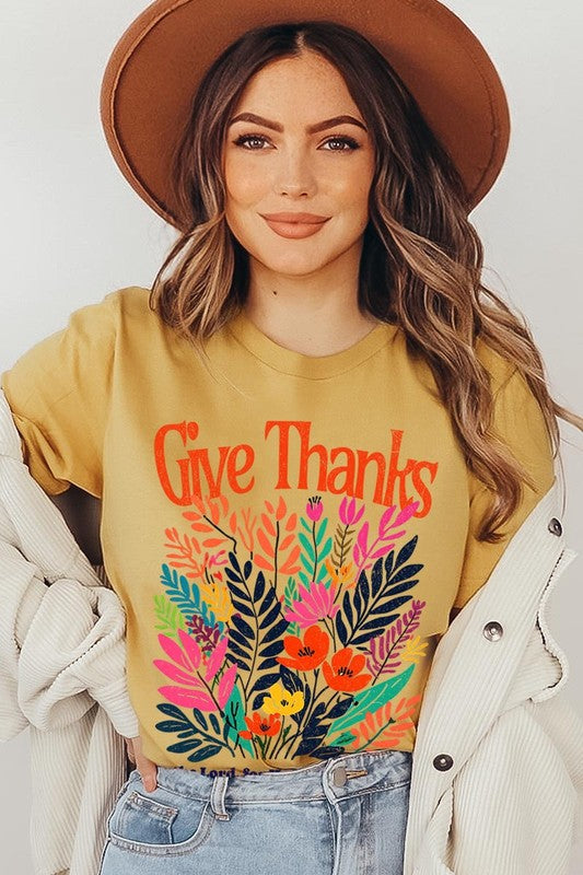 A person wears the Give Thanks Christian Graphic Tee, a high-quality printed design white t-shirt featuring colorful floral graphics and the text "Give Thanks. To the Lord, for He is good; His love endures forever. 1 Chronicles 16:34." Perfect as a family group uniform or a Christian graphic tee for gatherings.