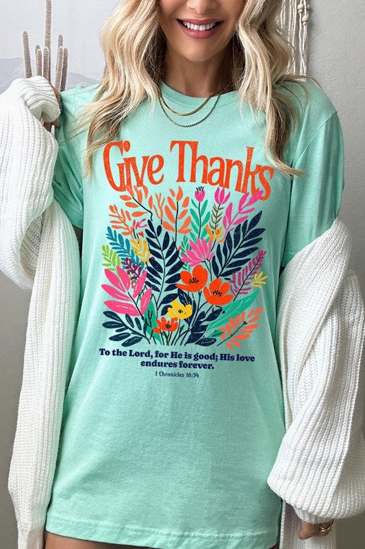 A person wears the Give Thanks Christian Graphic Tee, a high-quality printed design white t-shirt featuring colorful floral graphics and the text "Give Thanks. To the Lord, for He is good; His love endures forever. 1 Chronicles 16:34." Perfect as a family group uniform or a Christian graphic tee for gatherings.