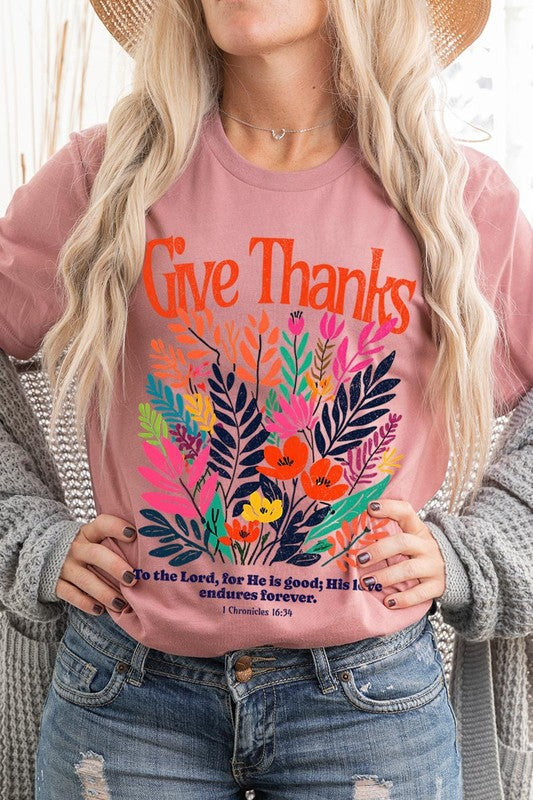 A person wears the Give Thanks Christian Graphic Tee, a high-quality printed design white t-shirt featuring colorful floral graphics and the text "Give Thanks. To the Lord, for He is good; His love endures forever. 1 Chronicles 16:34." Perfect as a family group uniform or a Christian graphic tee for gatherings.