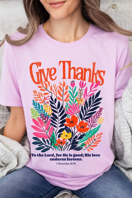 A person wears the Give Thanks Christian Graphic Tee, a high-quality printed design white t-shirt featuring colorful floral graphics and the text "Give Thanks. To the Lord, for He is good; His love endures forever. 1 Chronicles 16:34." Perfect as a family group uniform or a Christian graphic tee for gatherings.