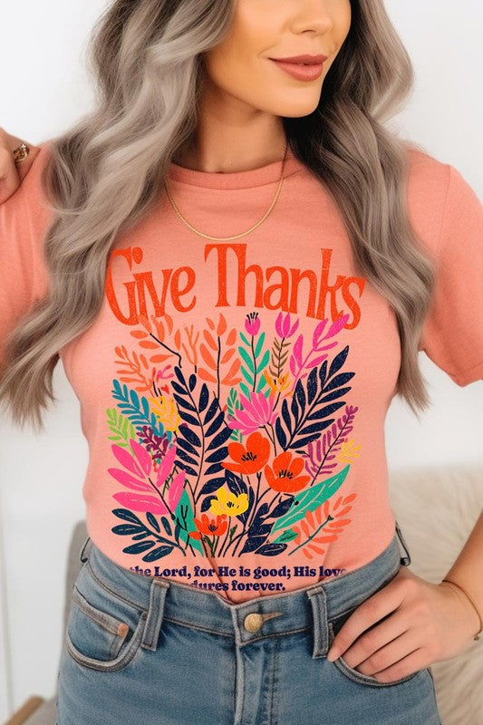 A person wears the Give Thanks Christian Graphic Tee, a high-quality printed design white t-shirt featuring colorful floral graphics and the text "Give Thanks. To the Lord, for He is good; His love endures forever. 1 Chronicles 16:34." Perfect as a family group uniform or a Christian graphic tee for gatherings.