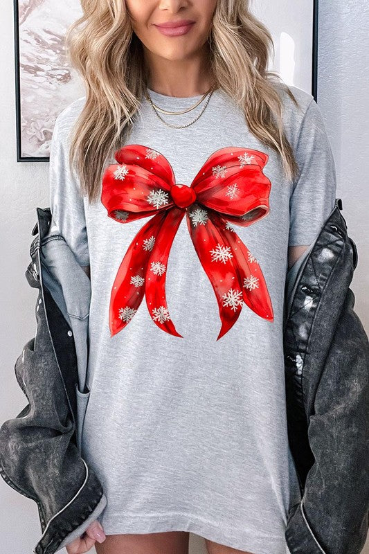 A woman wearing a Christmas Snowflakes Bow Graphic Tee in beige, featuring a large red bow design. She has long hair and is holding a red and black plaid shirt.