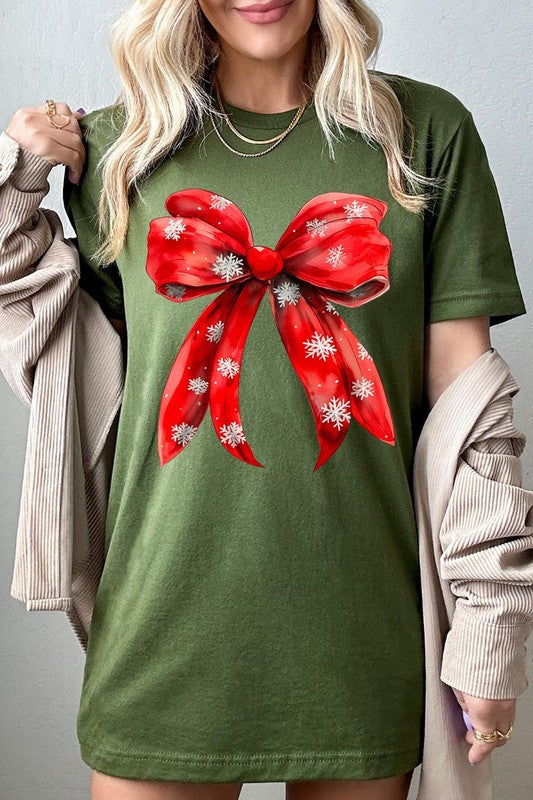 A woman wearing a Christmas Snowflakes Bow Graphic Tee in beige, featuring a large red bow design. She has long hair and is holding a red and black plaid shirt.