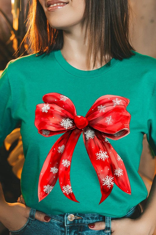 A woman wearing a Christmas Snowflakes Bow Graphic Tee in beige, featuring a large red bow design. She has long hair and is holding a red and black plaid shirt.