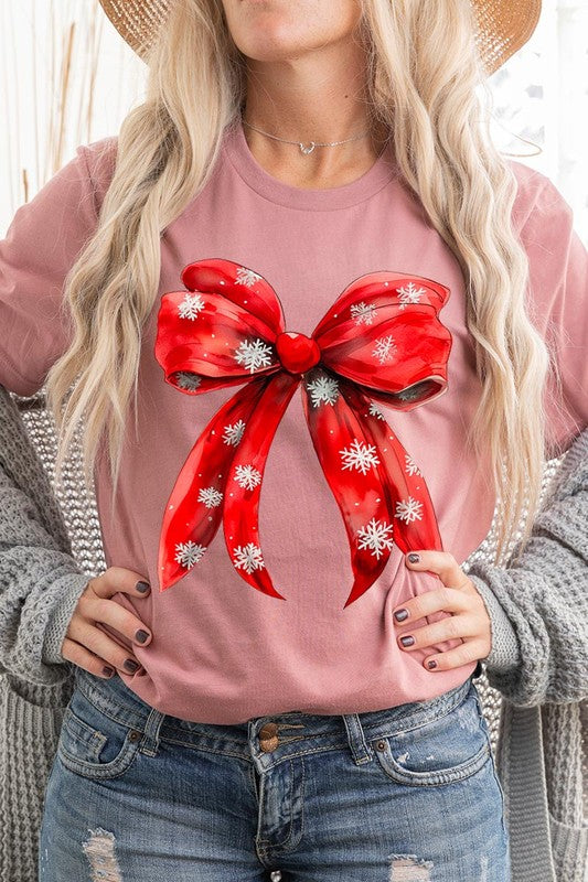 A woman wearing a Christmas Snowflakes Bow Graphic Tee in beige, featuring a large red bow design. She has long hair and is holding a red and black plaid shirt.