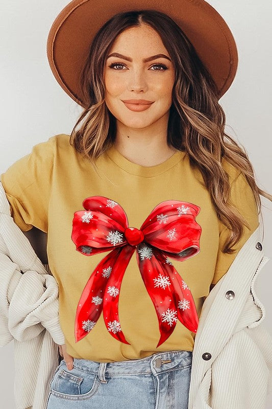 A woman wearing a Christmas Snowflakes Bow Graphic Tee in beige, featuring a large red bow design. She has long hair and is holding a red and black plaid shirt.