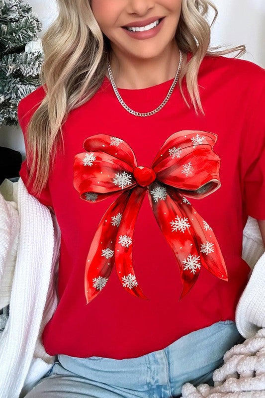 A woman wearing a Christmas Snowflakes Bow Graphic Tee in beige, featuring a large red bow design. She has long hair and is holding a red and black plaid shirt.