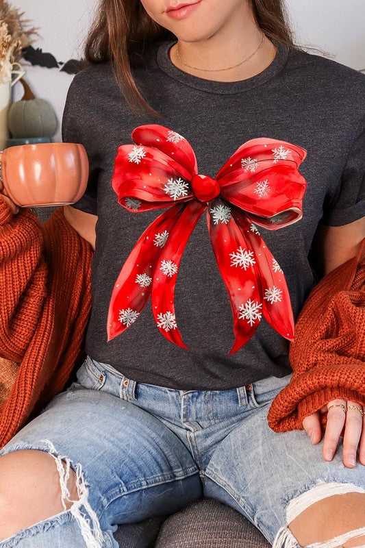 A woman wearing a Christmas Snowflakes Bow Graphic Tee in beige, featuring a large red bow design. She has long hair and is holding a red and black plaid shirt.