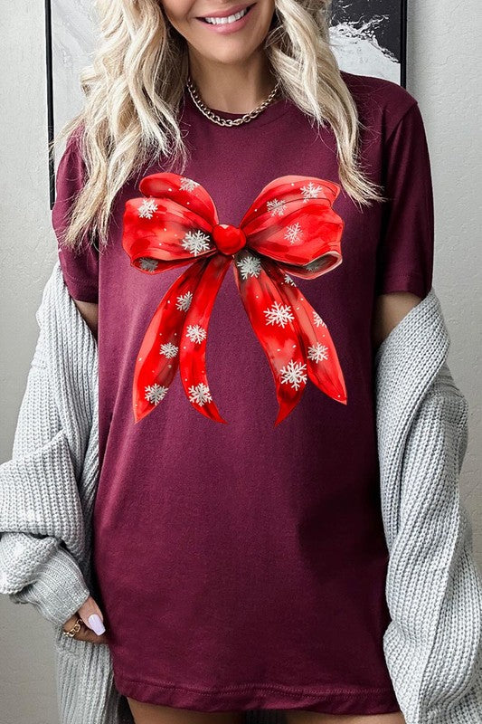 A woman wearing a Christmas Snowflakes Bow Graphic Tee in beige, featuring a large red bow design. She has long hair and is holding a red and black plaid shirt.