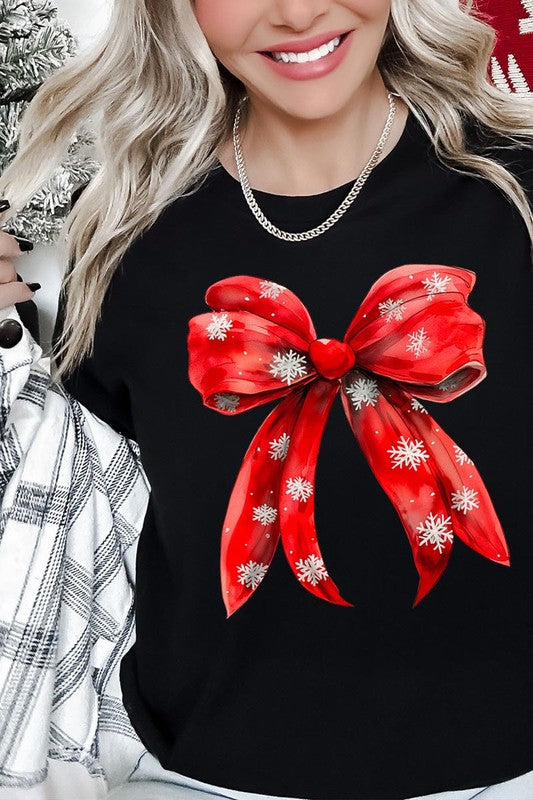 A woman wearing a Christmas Snowflakes Bow Graphic Tee in beige, featuring a large red bow design. She has long hair and is holding a red and black plaid shirt.