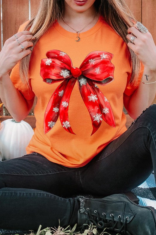 A woman wearing a Christmas Snowflakes Bow Graphic Tee in beige, featuring a large red bow design. She has long hair and is holding a red and black plaid shirt.