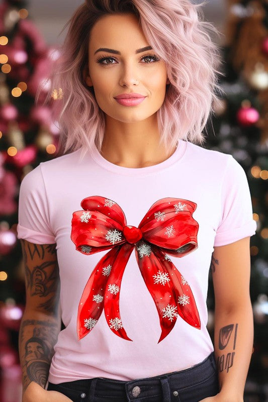 A woman wearing a Christmas Snowflakes Bow Graphic Tee in beige, featuring a large red bow design. She has long hair and is holding a red and black plaid shirt.