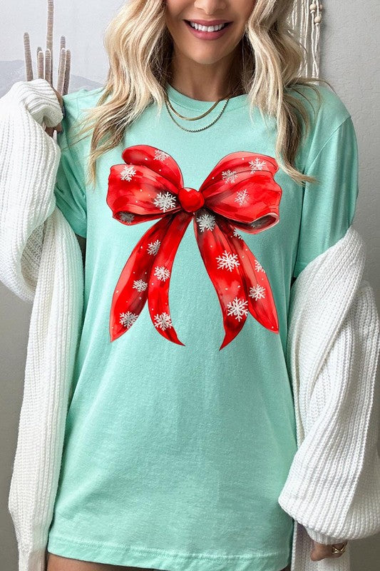 A woman wearing a Christmas Snowflakes Bow Graphic Tee in beige, featuring a large red bow design. She has long hair and is holding a red and black plaid shirt.