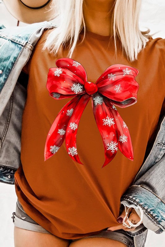 A woman wearing a Christmas Snowflakes Bow Graphic Tee in beige, featuring a large red bow design. She has long hair and is holding a red and black plaid shirt.