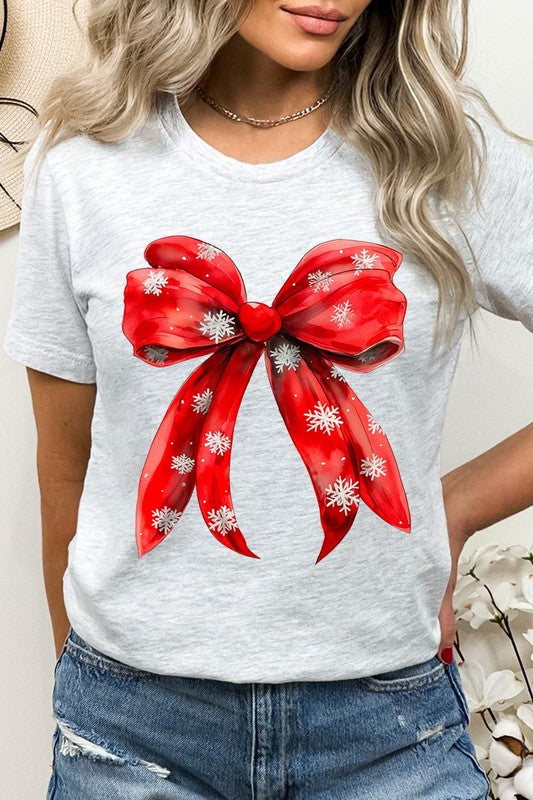 A woman wearing a Christmas Snowflakes Bow Graphic Tee in beige, featuring a large red bow design. She has long hair and is holding a red and black plaid shirt.