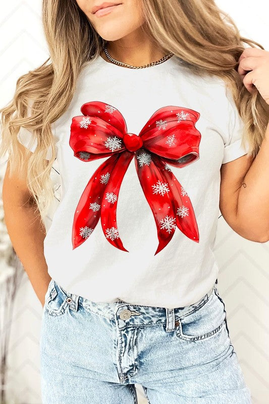 A woman wearing a Christmas Snowflakes Bow Graphic Tee in beige, featuring a large red bow design. She has long hair and is holding a red and black plaid shirt.