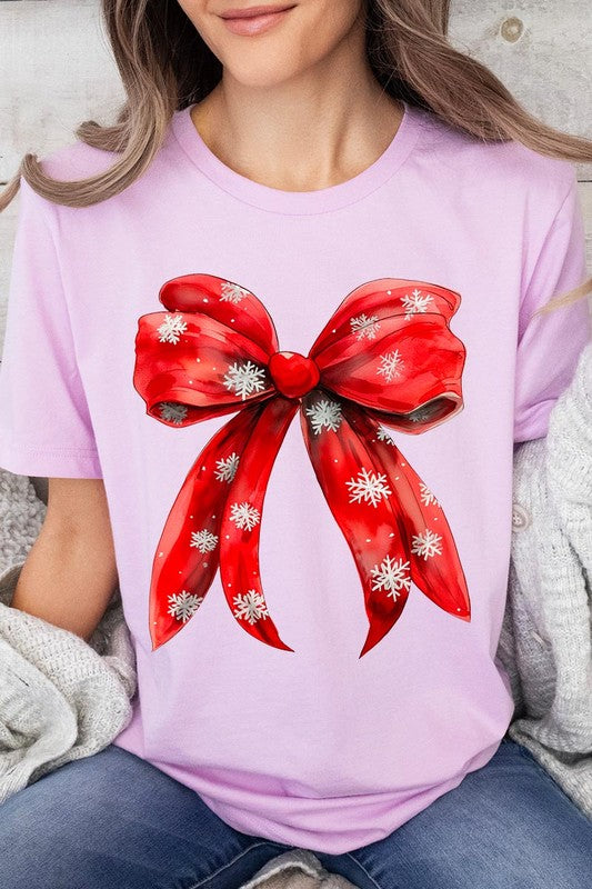 A woman wearing a Christmas Snowflakes Bow Graphic Tee in beige, featuring a large red bow design. She has long hair and is holding a red and black plaid shirt.