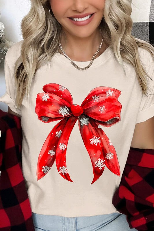 A woman wearing a Christmas Snowflakes Bow Graphic Tee in beige, featuring a large red bow design. She has long hair and is holding a red and black plaid shirt.