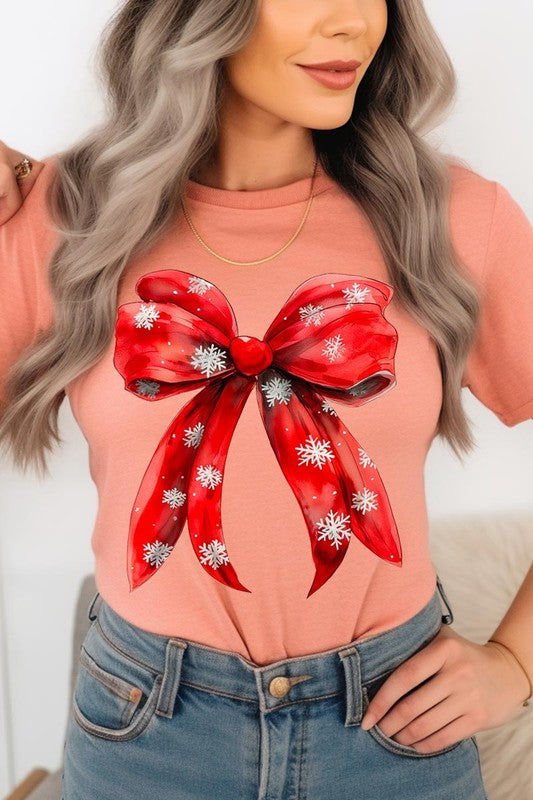 A woman wearing a Christmas Snowflakes Bow Graphic Tee in beige, featuring a large red bow design. She has long hair and is holding a red and black plaid shirt.