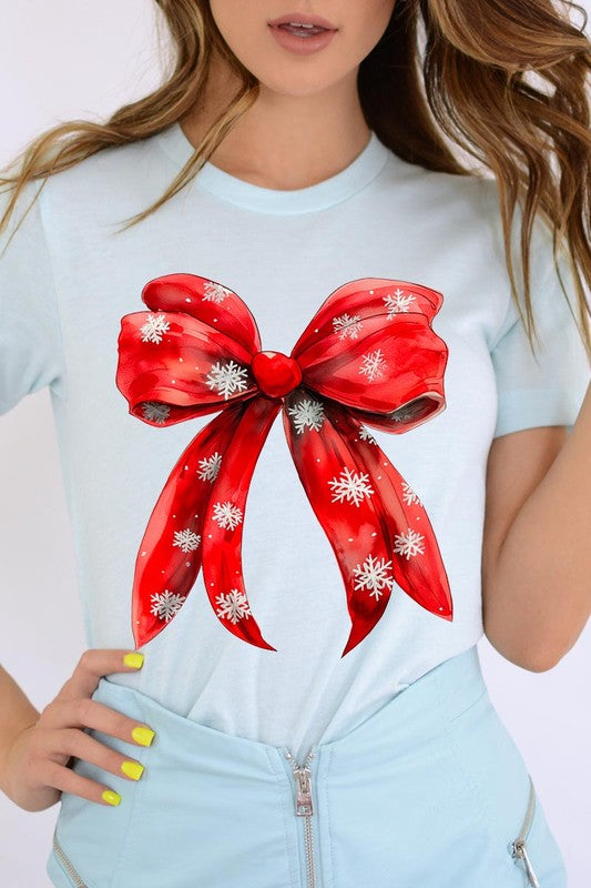 A woman wearing a Christmas Snowflakes Bow Graphic Tee in beige, featuring a large red bow design. She has long hair and is holding a red and black plaid shirt.