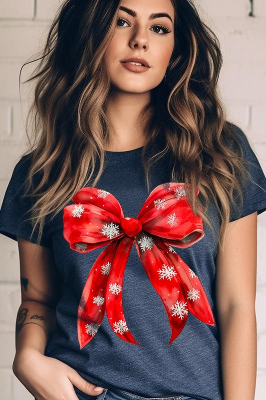 A woman wearing a Christmas Snowflakes Bow Graphic Tee in beige, featuring a large red bow design. She has long hair and is holding a red and black plaid shirt.