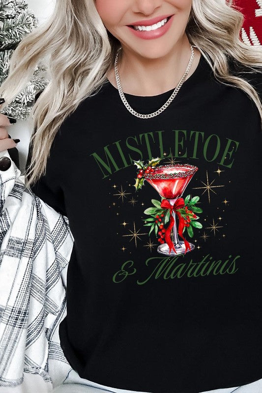 A person wearing the high-quality "Mistletoe & Martinis Graphic Tee" that features festive text and graphics, including a martini glass illustration adorned with holiday decorations, paired with a green plaid shirt.