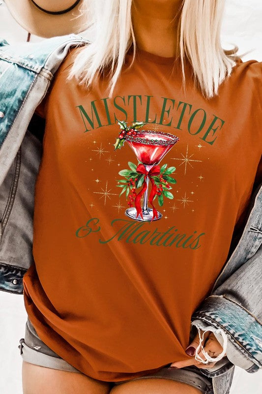 A person wearing the high-quality "Mistletoe & Martinis Graphic Tee" that features festive text and graphics, including a martini glass illustration adorned with holiday decorations, paired with a green plaid shirt.