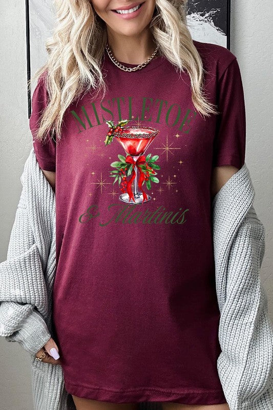 A person wearing the high-quality "Mistletoe & Martinis Graphic Tee" that features festive text and graphics, including a martini glass illustration adorned with holiday decorations, paired with a green plaid shirt.