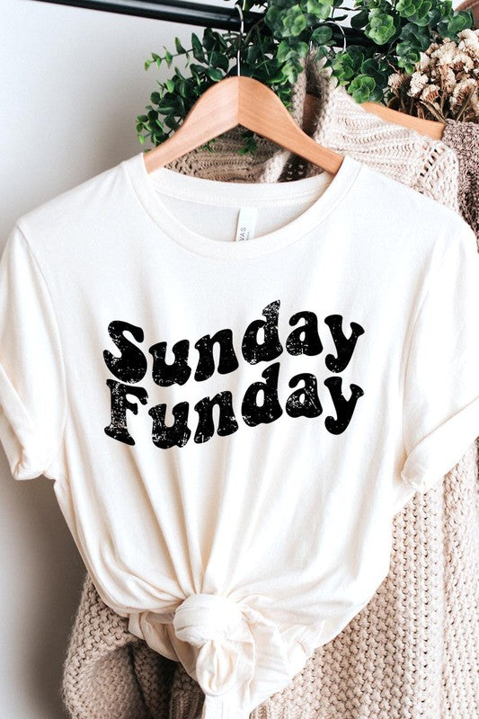 The "Sunday Funday" graphic tee with high-quality direct-to-film printing is paired with light blue distressed denim shorts, a brown hat, and a woven bag on a wooden hanger rack.