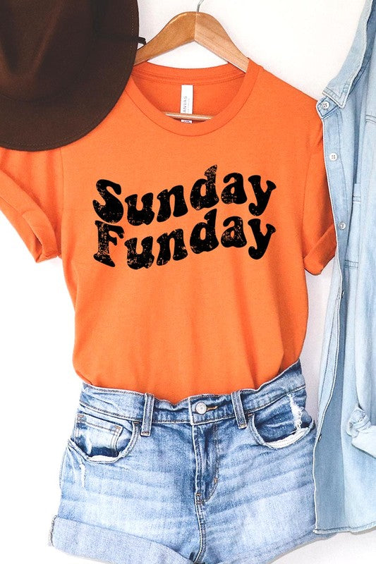 The "Sunday Funday" graphic tee with high-quality direct-to-film printing is paired with light blue distressed denim shorts, a brown hat, and a woven bag on a wooden hanger rack.