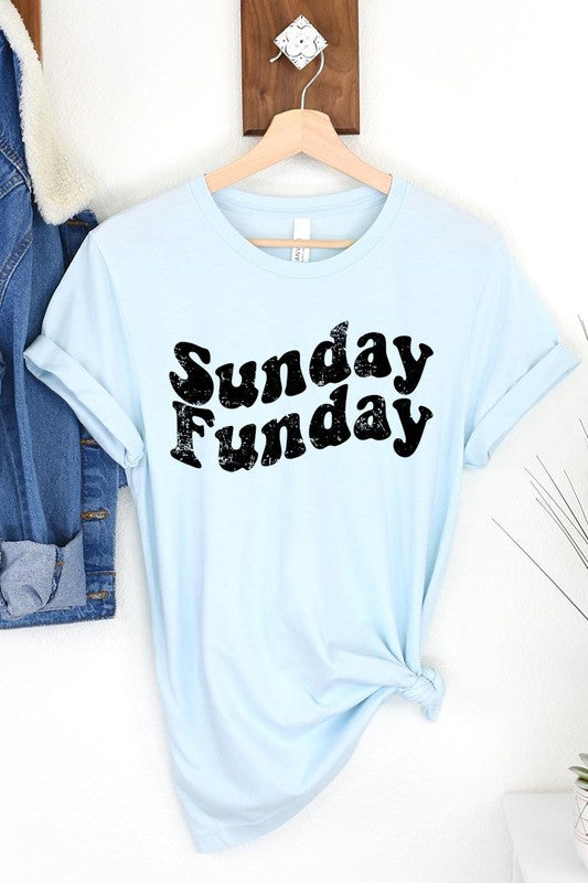 The "Sunday Funday" graphic tee with high-quality direct-to-film printing is paired with light blue distressed denim shorts, a brown hat, and a woven bag on a wooden hanger rack.