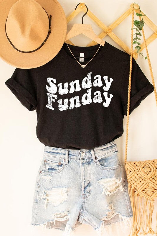 The "Sunday Funday" graphic tee with high-quality direct-to-film printing is paired with light blue distressed denim shorts, a brown hat, and a woven bag on a wooden hanger rack.