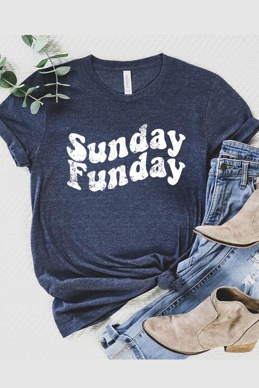 The "Sunday Funday" graphic tee with high-quality direct-to-film printing is paired with light blue distressed denim shorts, a brown hat, and a woven bag on a wooden hanger rack.