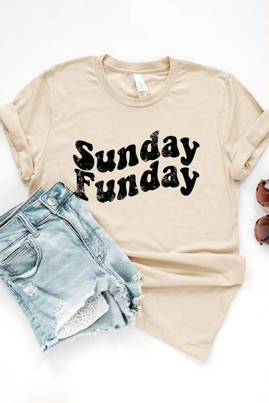 The "Sunday Funday" graphic tee with high-quality direct-to-film printing is paired with light blue distressed denim shorts, a brown hat, and a woven bag on a wooden hanger rack.