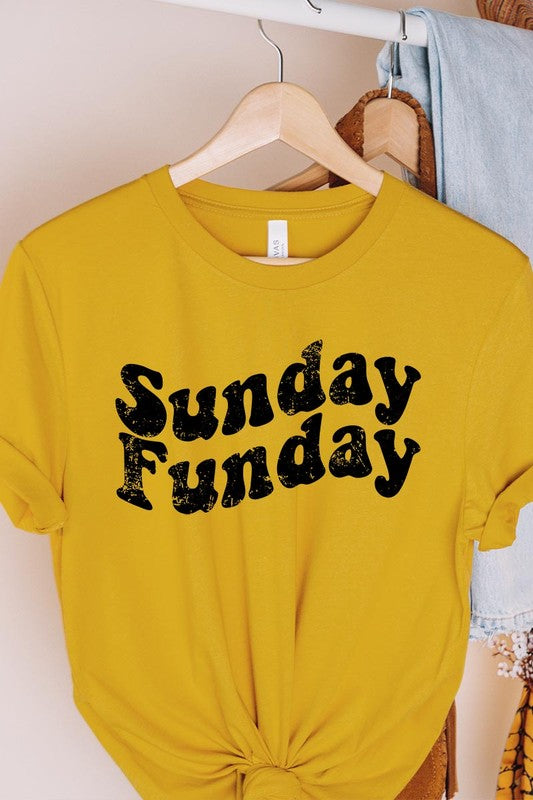 The "Sunday Funday" graphic tee with high-quality direct-to-film printing is paired with light blue distressed denim shorts, a brown hat, and a woven bag on a wooden hanger rack.