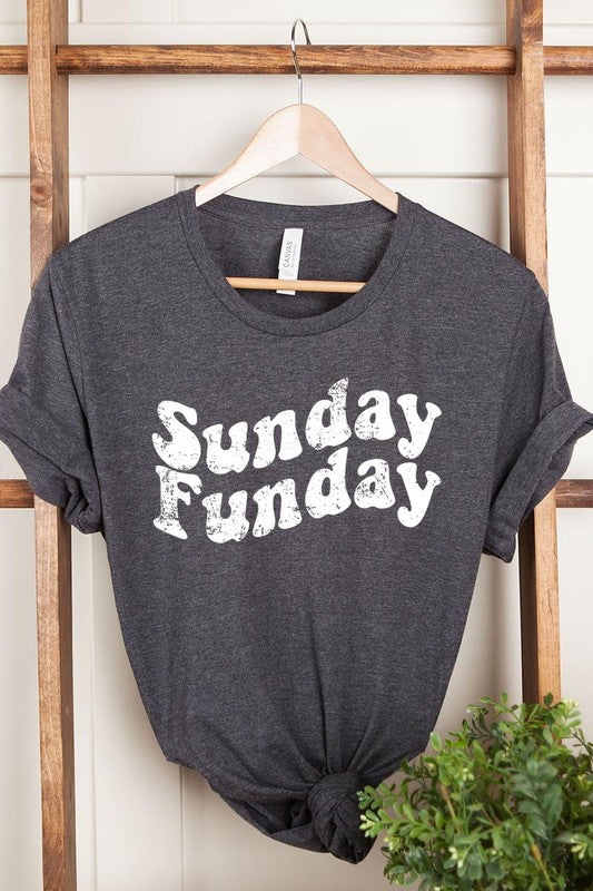 The "Sunday Funday" graphic tee with high-quality direct-to-film printing is paired with light blue distressed denim shorts, a brown hat, and a woven bag on a wooden hanger rack.