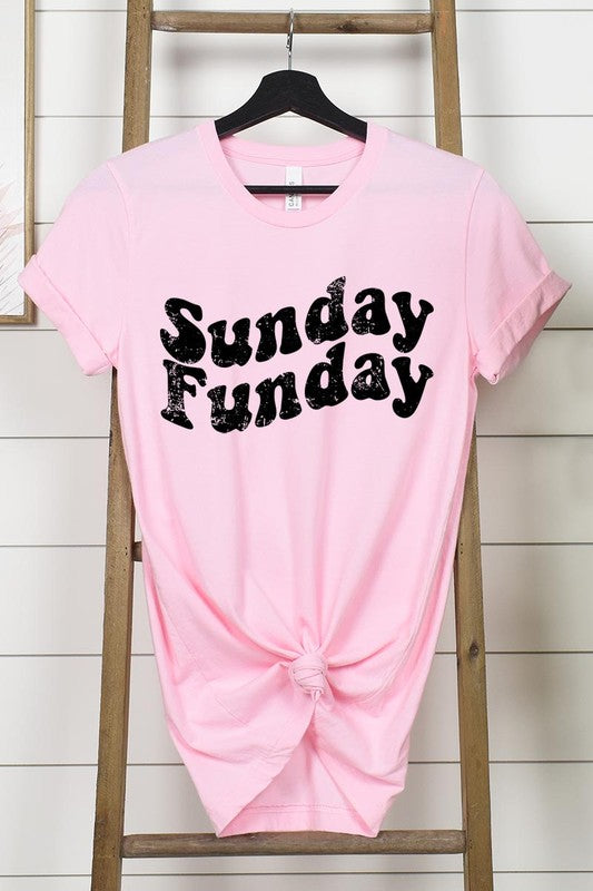 The "Sunday Funday" graphic tee with high-quality direct-to-film printing is paired with light blue distressed denim shorts, a brown hat, and a woven bag on a wooden hanger rack.