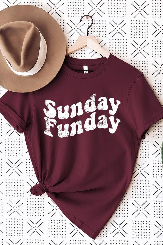 The "Sunday Funday" graphic tee with high-quality direct-to-film printing is paired with light blue distressed denim shorts, a brown hat, and a woven bag on a wooden hanger rack.