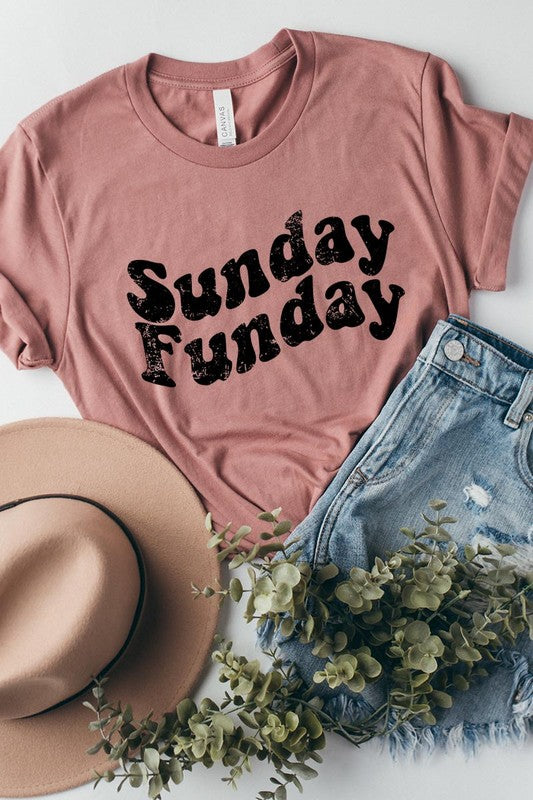 The "Sunday Funday" graphic tee with high-quality direct-to-film printing is paired with light blue distressed denim shorts, a brown hat, and a woven bag on a wooden hanger rack.