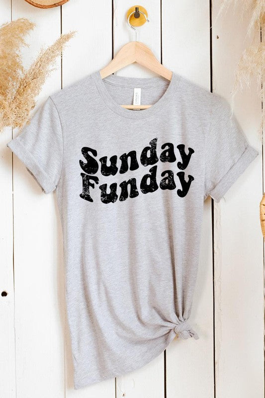 The "Sunday Funday" graphic tee with high-quality direct-to-film printing is paired with light blue distressed denim shorts, a brown hat, and a woven bag on a wooden hanger rack.